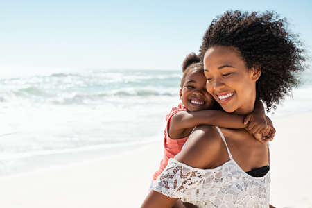 Smiling young black mother and beautiful daughter having fun on the beach with copy space. Portrait of happy sister giving a piggyback ride to cute little girl at seaside. Lovely female kid embracing her lovely mom during summer vacation.の写真素材