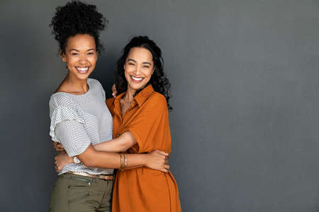 Portrait of happy women embracing each other against grey wall with copy space. Carefree latin woman hugging her best friend while standing against gray background while looking at camera. Smiling lesbian couple having fun and laughing together.の写真素材