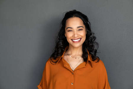 Portrait of latin young woman with pleasant smile isolated on grey wall with copy space. Carefree hispanic woman smiling and looking at camera standing on gray background. Beautiful multiethnic girl in casual clothing laughing against grey wall.の写真素材