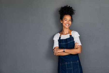 Young black woman wearing denim apron isolated on grey background with copy space. Portrait of successful african american woman with crossed arms on gray wall. Smiling black waitress and cafe owner standing against grey wall while looking at camera.の写真素材