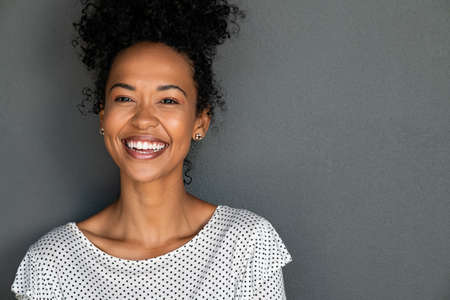 Portrait of cheerful young woman laughing against grey background. Happy african american girl with curly hair smiling against gray wall with copy space. Close up face of young black woman smiling.の写真素材