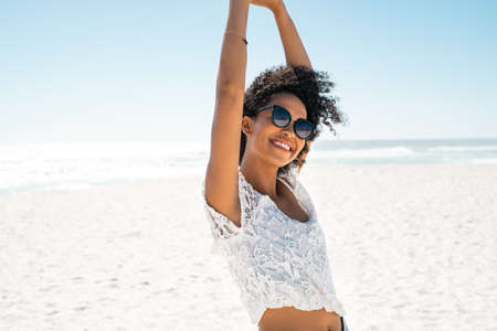Smiling young african american woman stretching arms while relaxing at beach. Beautiful black woman dancing at seaside during weekend while looking at camera. Happy carefree girl with afro hair wearing sunglasses with copy space: freedom and positive vibrations.の写真素材