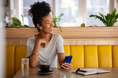 Smiling young woman with earphones holding phone and drinking coffee in modern cafe while looking away. Cheerful black woman using mobile phone and wireless earphones while doing a call in cafe. Positive beautiful girl sitting and relaxing in coffee shop.の写真素材