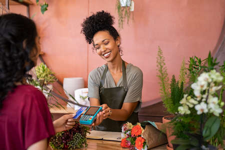 Smiling and friendly florist holding card reader machine at counter with customer paying with credit card. Young african american florist shop assistant holding payment machine while buyer purchase a bunch flower. Woman using bank credit card to make payment by NFC machine.の写真素材