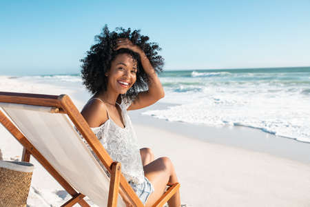Portrait of happy african american woman relaxing on wooden deck chair at  tropical beach while looking at camera. Smiling black girl enjoying vacation at seaside. Beautiful woman sunbathing and relaxing at sea with copy space.の写真素材