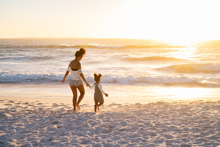 Happy african american mother relaxing with cute little girl at beach while walking towards the sea. Black family walking towards ocean on beach during sunset with copy space. Mother and child running barefoot on seaside while holding hands.の写真素材