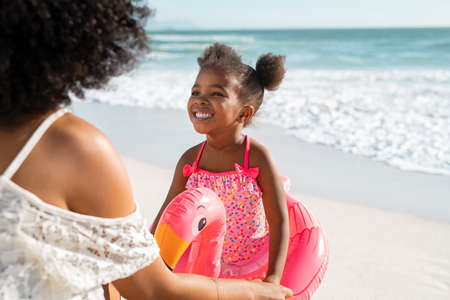 Rear view of young african american mother holding hands and talking to daughter standing with float for swim at beach. Happy young woman palying with female child inside flamingo inflatable toy. Mom and female kid having fun during summer vacation at sea.の写真素材