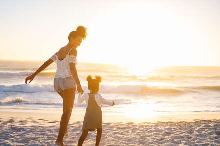Smiling african american mother walking with cute little girl at beach. Lovely black woman holding hands with little daughter while walking towards ocean at beach during sunset. Mother and child running barefoot and jumping on beach as the sun sets with copy space.の写真素材