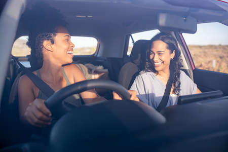 Young beautiful friends laughing while traveling in car during a road trip. Two multiethnic women driving in car and having fun. Two attractive girls talking together while driving in a car through the countryside on a sunset.の写真素材