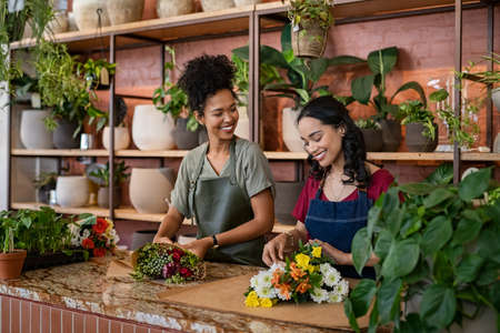 Two happy and smiling young woman florist working together in flower shop. Female florist talking and making beautiful bouquet of flower together with her black colleague. Beautiful florists arranging bunch of flower at counter at floral shop.の写真素材