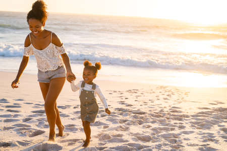 Young beautiful woman with her adorable little cute girl running on tropical beach during sunset. Cheerful and cute african american daughter walking and playing with mother on beach together. Lovely bigger sister and female child enjoying beach with copy space.の写真素材