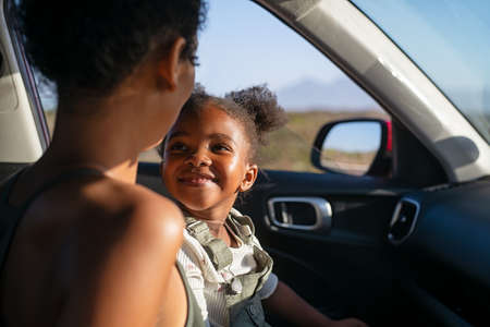 Cute black little girl looking her mother while sitting in car during sunset. Cheerful mother and daughter sitting in car during road trip. Smiling and carefree female child laughing with african american mother.の写真素材