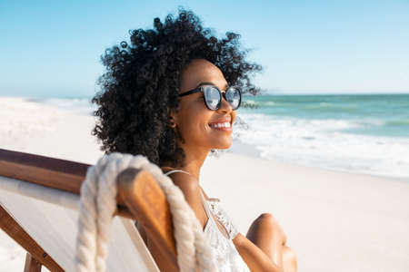 Portrait of happy young black woman relaxing on deck chair at beach wearing spectacles. Smiling african american girl with sunglasses enjoy vacation at beach. Carefree happy young woman sunbathing and relaxing at sea with copy space.の写真素材