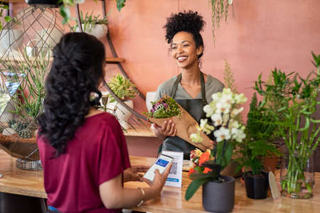 Young african florist holding beautiful bouquet of fresh flower while customer making online payment with smartphone while scanning QR code. Black saleswoman wearing apron giving a bunch of flower to customer at botany shop. Young woman making digital payment by phone wallet at flower shop.の写真素材