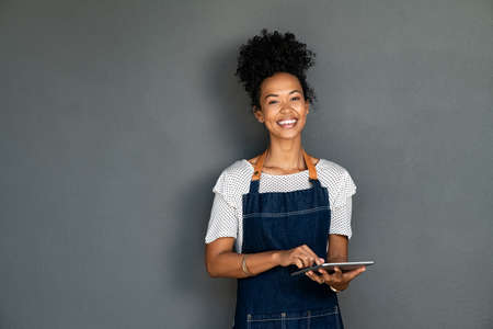 Portrait of beautiful young cafe black waitress using digital tablet on grey background. Happy young black woman in blue jeans apron working on digital tablet while looking at camera. Successful african american cafe owner isolated against gray wall with copy space.の写真素材