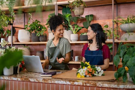 Two women in florist shop working on laptop and preparing online order of bouquet of flower. Smiling two colleagues florists arranging bunch of flowers and using laptop while working at flower shop. Young florist using laptop and preparing flower order online while having a discussion with colleague.の写真素材