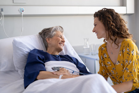 Young daughter visiting sick old mother in hospital. Happy senior patient smiling while in conversation with her lovely daughter. Granddaughter visiting and cheering her hospitalized grandmother lying in bed at hospital ward after surgery.の写真素材