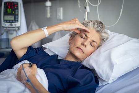 Senior woman suffering from headache while lying on hospital with eyes closed and drip in hand. Sad woman with grey hair in private clinic feeling sick. Recovering depressed mature woman in modern hospital ward.の写真素材