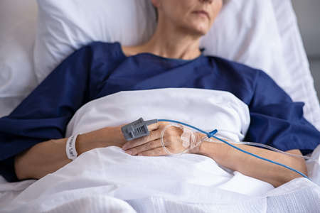 Close up of senior woman lying on bed with saline solution tube intravenous drip in hand. Mature patient hospitalized with IV drip in hand while undergoing treatment at hospital. A pulse oximeter used to measure pulse rate and oxygen levels on old patient.の写真素材