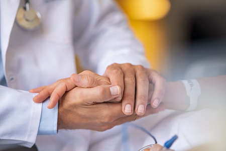 Close up of doctor hands holding senior patient's hands and comforting her. Close up of general practitioner holding senior woman hand giving assurance and comfort. Doctor comforting patient at consulting room. Help, support and caring concept.の写真素材