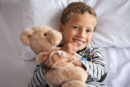 Happy little boy hugging teddy bear with drip in hand smiling on hospital bed. Portrait of smiling child embracing teddy bear on gurney at private clinic. Cute kid with stuff toy lying on hospital bed and looking at camera with big grin.の写真素材