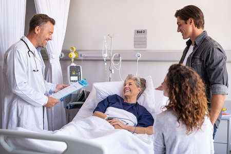 Mature happy doctor holding clipboard giving good news to family and old patient. Smiling senior woman lying on hospital bed with son and daughter visiting and talking to physician. Doctor visiting hospitalized patient with family in modern hospital ward.の写真素材