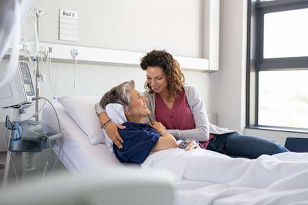 Lovely young daughter visiting old sick mother hospitalized in private clinic. Reassuring woman assisting her senior ill mother lying on hospital bed and talking. Senior and parent care concept.の写真素材
