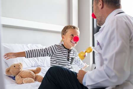 Smiling little boy wearing red nose and flying with outstreched arms like superhero in hospital bed with doctor. Happy playful child playing and talking to funny pediatrician at private clinic. Kid joking during medical examination with physician, clown therapy concept.の写真素材
