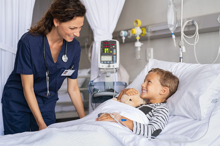 Happy young boy lying on gurney talking to friendly nurse. Smiling healthcare worker taking care of cute little boy lying on bed in hospital with IV drip. Paramedic tuck the covers back to the patient in pediatrics ward in private clinic.の写真素材
