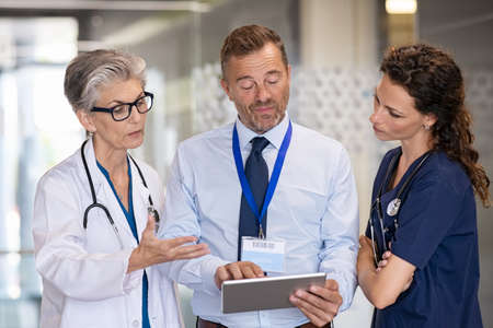 Senior woman doctor having a discussion in hospital hallway with medical staff. Doctor discussing patient case status with his team after operation. Pharmaceutical representative showing medical report on digital tablet about new medicine or therapy.の写真素材