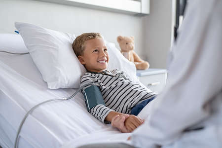 Smiling young boy lying on hospital bed getting blood pressure checked. Good child patient looking at doctor while holding hand during check up at private clinic. Strong happy kid with his teddy bear recovering from sickness in clinic, lying on gurney.の写真素材