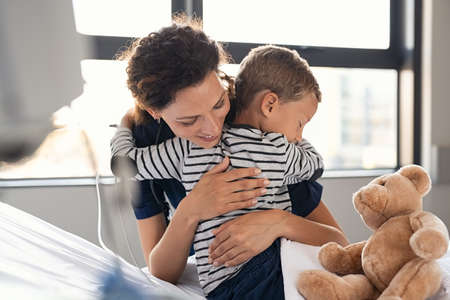 Happy nurse hugging sick little boy patient in hospital. Young medical practitioner embracing upset kid in clinic. Lovely doctor taking care of cute child at private clinic: gratitude and healthcare concept.の写真素材
