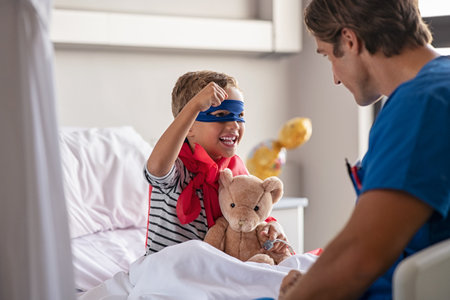 Strong little boy wearing superhero blue mask and red cape sitting on hospital bed playing with nurse. Happy superhero kid gesturing with playful doctor at clinic. Happy child playing as superman in pediatric ward with healthcare worker.の写真素材