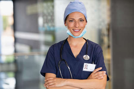 Portrait of  successfurl young woman doctor wearing face mask and scrubs in hospital. Confident and reliable medical practitioner with surgical mask looking at camera. Smiling nurse standing in clinic hallway with crossed arms and copy space.の写真素材