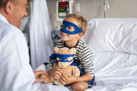 Cute little boy with superhero mask embracing teddy bear while playing with doctor and sitting on hospital bed. Playful child dressed as superhero holding stuff toy talking to pediatrician. Mature doctor visiting his little patient in hospital.の写真素材