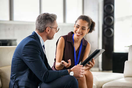 Young black businesswoman discussing work in office using digital tablet. African american woman manager in conversation with executive during conference. Professional mature businessman and smiling happy woman working while sitting on couch and discussing.の写真素材