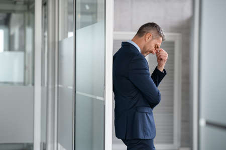 Tired business man suffering from headache at work. Mid adult entrepreneur feeling stressed while pressing eye standing and leaning at glass wall in modern office. Depressed businessman suffering from migraine while trying to work in corporate with copy space.の写真素材