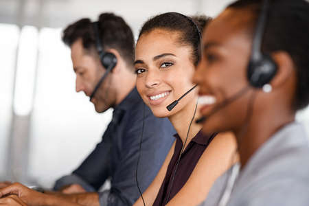 Portrait of smiling customer service operator with headset sitting with colleagues and attending calls in office. Beautiful black woman telemarketing agent working in call center with team and looking at camera. Smiling african american customer service representative sitting in a row.の写真素材