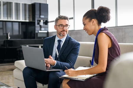 Mature businessman discussing work with young businesswoman while she is writing notes. Young black woman secretary writing notes on diary while man executive showing project on laptop. Professional man and african american woman discussing work during conference.の写真素材