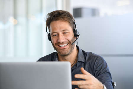 Technical support operator working with headset in office. Smiling handsome man working as call centre operator, speaking to customer and using laptop. Happy successful businessman working remotely while doing video conference.の写真素材