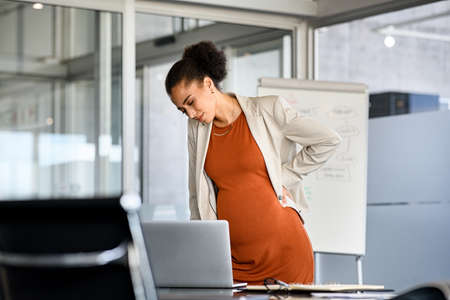 Young pregnant african american woman suffering from pain while working in office. Black pregnant woman feeling sick and tired at office while working on laptop. Woman entrepreneur suffering from back pain during pregnancy in modern office.の写真素材