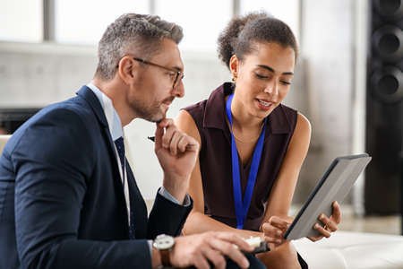 Young business woman discussing work with manager in office. Young black woman secretary showing documents on digital tablet to boss. Mature businessman and secretary working on office sitting on couch and discussing on digital report.の写真素材