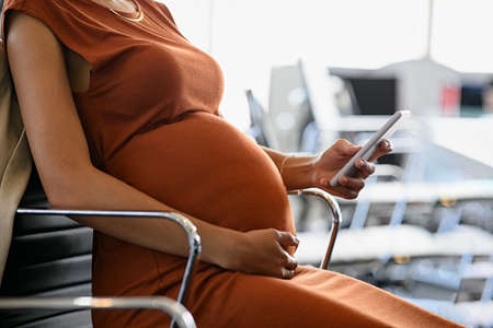 Close up hand of african pregnant woman holding belly while using smartphone in office. Pregnant young businesswoman writing email on mobile phone while working. Expecting woman reading text message on smart phone while sitting at desk.の写真素材