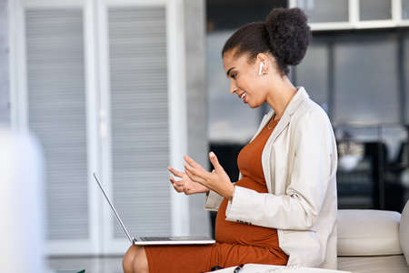 Smiling business woman talking to customer on video call using laptop with earphones. Young pregnant woman entrepreneur working online in a meeting while using computer in office. Pregnant african american businesswoman in conversation with colleague online using laptop.の写真素材