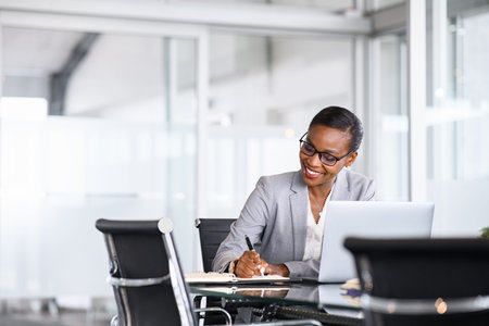 Mature businesswoman taking notes in notebook while using laptop at office. Mid adult black woman entrepreneur writing details on book while working on laptop in modern office. Smiling african american business woman with spectacles writing schedule and planning during video call.の写真素材
