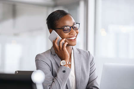 Mid adult businesswoman talking on cellphone in office. Smiling african american business woman in happy conversation on mobile phone. Mature confident black woman entrepreneur sitting at table and working from office.の写真素材