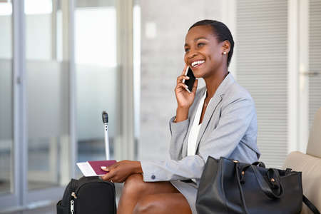 Smiling african businesswoman in formal clothing sitting in waiting room and talking over smartphone. Mature black beautiful woman traveller talking on cell phone at airport lounge. Successful woman on mobile phone at terminal airport.の写真素材