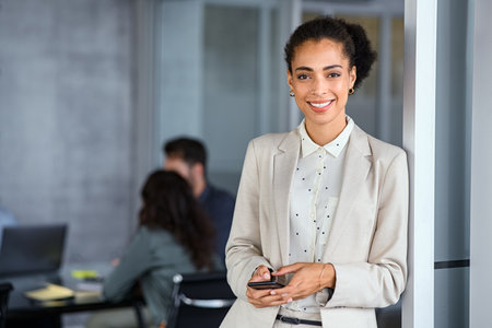 Portrait of beautiful businesswoman writing a text on smart phone with copy space in modern office. Smiling and proud black woman reading a new message on smartphone. Confident young african american business woman checking email at office while looking at camera.の写真素材