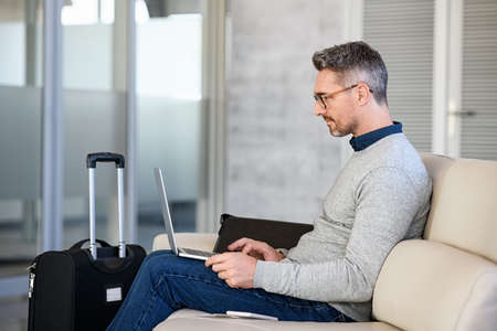 Successful mature man working on computer at airport in waiting room. Mid adult business man with luggage in international airport working on laptop while waiting for flight. Casual businessman passenger sitting on sofa in terminal hall and send an email on laptop.の写真素材