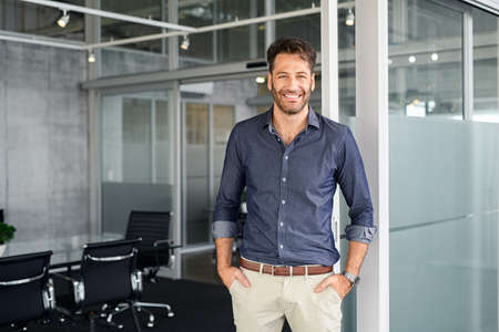Portrait of mature successful man with hands in pocket standing outside office conference room. Handsome businessman standing in office while looking at camera. Young satisfied man in formalwear standing in boardroom with copy space.の写真素材